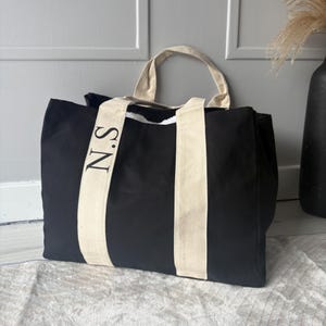 May include: Black tote bag with cream-coloured handles and vertical stripes. The bag features the initials "N.S" printed in black on one of the cream stripes. The bag is sitting on a light-coloured surface.