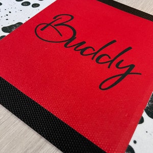 May include: Red doormat with a black border and a white and black dalmatian print. The doormat has the word "Buddy" written in black cursive.