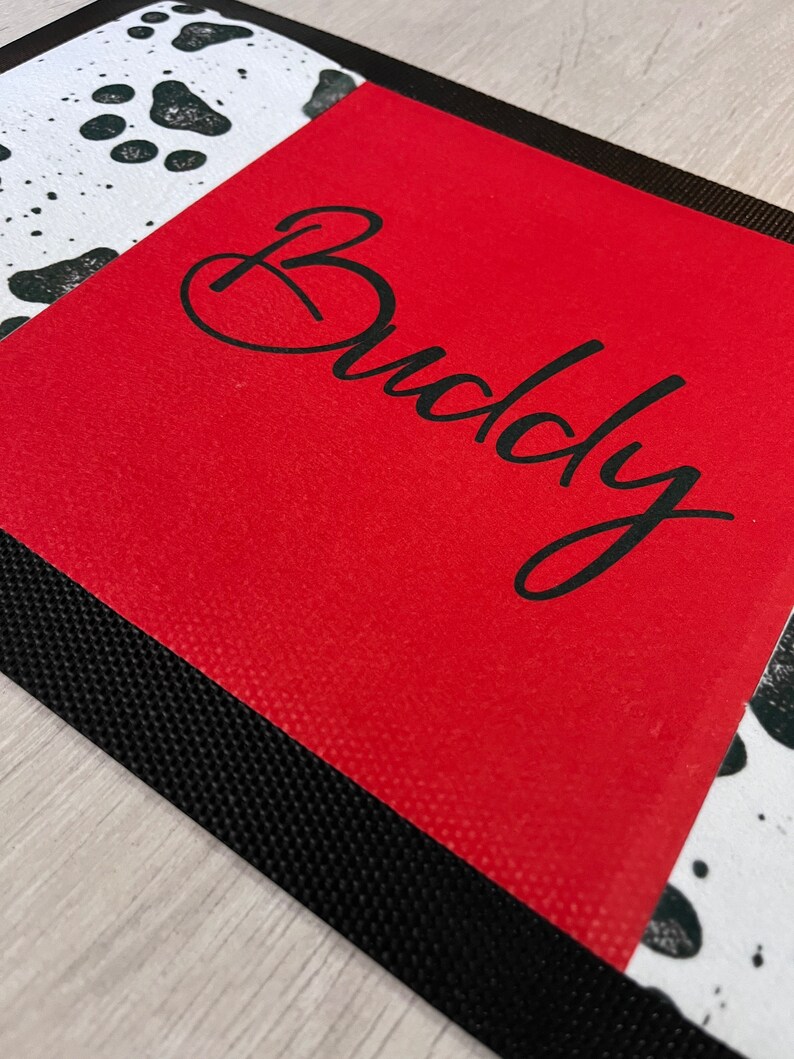 May include: Red doormat with a black border and a white and black dalmatian print. The doormat has the word "Buddy" written in black cursive.