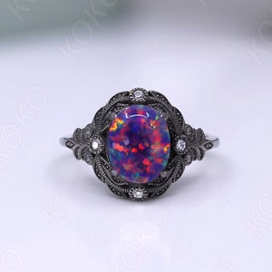 May include: A black ring with a large oval opal gemstone surrounded by smaller clear gemstones. The ring has a detailed floral design.