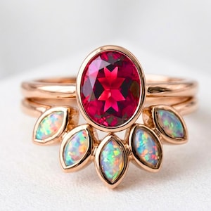 May include: A rose gold ring set featuring a central oval-cut deep red gemstone in a bezel setting. The band is complemented by a curved guard ring adorned with five marquise-cut iridescent opals arranged in a fan-like pattern along the base.