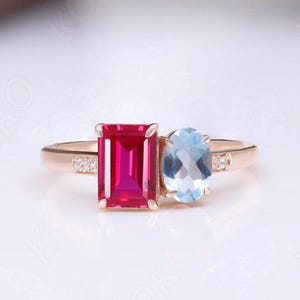 May include: A rose gold ring featuring a rectangular, vibrant red gemstone and an oval, light blue gemstone. Small accent stones are set on either side of the ring's band.