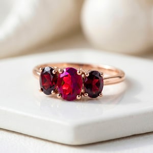 Può includere: Anello in oro rosa con tre pietre preziose ovali. La pietra centrale è magenta, affiancata da due pietre rosso scuro. L'anello è appoggiato su una superficie bianca, con uno sfondo sfocato.