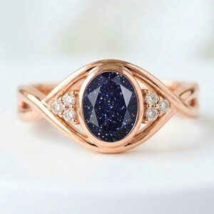 May include: A rose gold ring featuring a central oval-cut dark blue sandstone with sparkling inclusions. The band has a twisted design and is accented on both sides of the center stone by three small, clear, round-cut stones set in a triangular pattern.
