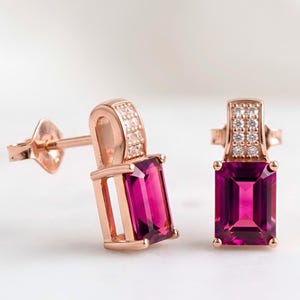 May include: A pair of rose gold earrings, each with a rectangular, deep pink gemstone. The earrings have a small, curved top section set with small, clear stones. They have a post and butterfly clasp.