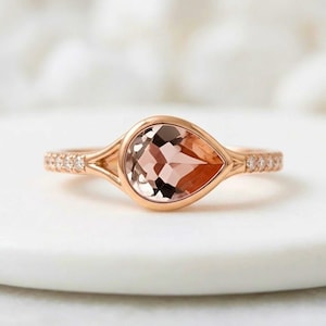 May include: A rose gold ring featuring a pear-shaped peach-colored gemstone set in a bezel setting. The band splits as it reaches the center stone and is accented with small round clear stones along the sides of the shank on both sides.