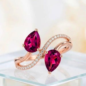 May include: A rose gold bypass ring featuring two pear-cut deep pink gemstones. The band is split into two curved sections, with one side encrusted with small, clear round-cut stones that follow the contour of the ring and frame the gems.