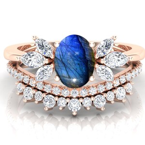 May include: A rose gold ring with a large oval blue labradorite gemstone surrounded by smaller clear gemstones. The ring has a delicate floral design with a row of smaller gemstones around the band.