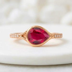 May include: A rose gold ring featuring a pear-shaped, deep red gemstone set in a bezel setting. The band splits into two delicate arms at the center and is accented with small, clear round stones embedded along both sides of the metal shank.