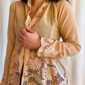 Puede incluir: Una blusa de color melocotón claro con intrincados detalles de encaje floral. La blusa presenta mangas largas y un cuello mandarín, con el ribete de encaje acentuando el escote, los puños y la abertura frontal. El bordado delicado añade un toque de elegancia.