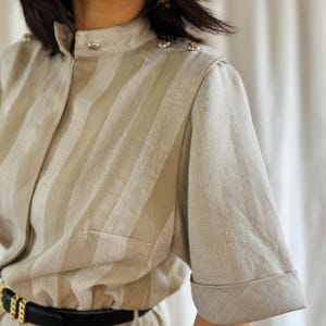 Könnte beinhalten: Beige-weiß gestreiftes Kleid mit kurzen Ärmeln und Stehkragen. Das Kleid hat silberne Knöpfe auf den Schultern und einen schwarzen Gürtel mit goldener Schnalle. Das Kleid hat eine taillierte Passform.