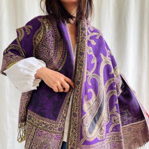 May include: A purple paisley patterned shawl with gold accents and fringe. The shawl is draped over a person's shoulders.
