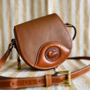 Könnte beinhalten: Braune Leder-Umhängetasche mit strukturierter Oberfläche. Die Tasche hat eine abgerundete Klappe, einen langen, verstellbaren Riemen und eine Messingschnalle. Das Markenlogo ist auf der Vorderseite sichtbar. Maße: ca. 20 cm breit und 18 cm hoch.