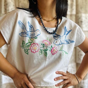 Op de afbeelding: Een wit shirt met korte mouwen voorzien van blauw geborduurde vogels en roze bloemaccenten met groene bladeren. Het kledingstuk heeft een ronde halslijn en wordt gedragen met een gouden ketting en een donkere kralenketting.