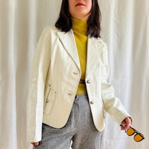 May include: A white leather jacket with a yellow turtleneck sweater underneath. The jacket has a button closure and two pockets on the front. The person is wearing gray pants.