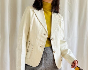 Könnte beinhalten: Eine weiße Lederjacke mit einem gelben Rollkragenpullover darunter. Die Jacke hat einen Knopfverschluss und zwei Taschen auf der Vorderseite. Die Person trägt eine graue Hose.