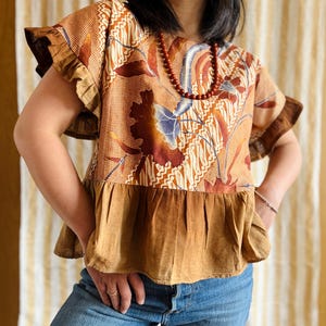 May include: A short-sleeved, brown batik top with a floral pattern and ruffled sleeves. The top features a contrasting brown peplum and is paired with blue jeans. A beaded necklace adds a touch of color.