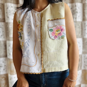 May include: A sleeveless, button-down vest with a patchwork design. The left side features a white floral pattern, while the right side is a light yellow grid pattern with a pocket. The pocket is adorned with a pink and yellow floral embroidery.