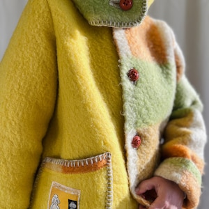 Könnte beinhalten: Ein gelber Wollmantel mit einem Patchwork-Design in Grün, Orange und Weiß. Der Mantel hat eine Kapuze, rote Knöpfe und eine aufgesetzte Tasche mit einem Etikett. Die sichtbaren Nähte tragen zur handgefertigten Ästhetik bei.