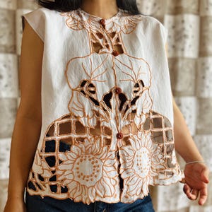 May include: White sleeveless top with intricate cut-out and embroidered floral and leaf designs in a warm brown color. The top features a button-up front and a high neckline. The design includes sunflower and grid patterns.
