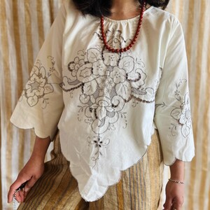 Op de afbeelding: Een crèmekleurige blouse met driekwartmouwen en een geschulpte zoom. De blouse heeft ingewikkelde bloemenborduursels in grijstinten en bruin over de voorkant en op de mouwen. Een rode kralenketting wordt bij de blouse gedragen.