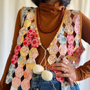 Op de afbeelding: Een patchwork vest met een kleurrijke reeks zeshoekige stoffen stukken in bloemen- en patroonontwerpen. Het vest heeft een gele rand, witte knopen en twee witte pompons. Gedragen over een bruine coltrui en een spijkerbroek.