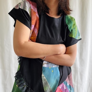 May include: Black short-sleeved top with a colourful patchwork design. The patchwork includes shades of green, blue, pink, and orange. The top has a black lace trim along the bottom edge.