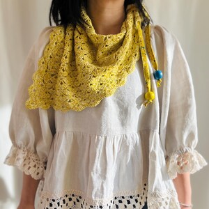 May include: A yellow crocheted shawl with a scalloped edge is draped over a white blouse. The shawl has a floral pattern and is secured with a yellow ribbon adorned with blue and yellow beads. The blouse has lace trim.