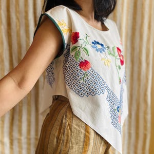 Op de afbeelding: Witte geborduurde top met een groene bies en bloemenmotief. De top heeft rode, blauwe en gele bloemen. De onderkant van de top heeft een blauw geometrisch patroon. Het model draagt een gestreepte broek.