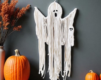 May include: A macrame ghost wall hanging with a large and small ghost design. The white ghost is hanging on a dark gray wall, with two small orange pumpkins and a large orange pumpkin. A vase with dried orange flowers is on the left.