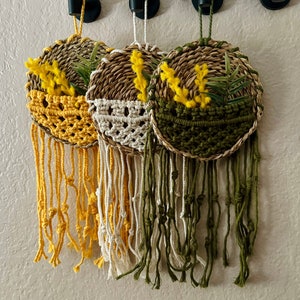 May include: Three macrame wall hangings with woven baskets and yellow flowers. The baskets are in shades of brown, cream, and green. The macrame is in matching colors with long fringe.