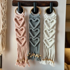 May include: Three macrame wall hangings with heart designs. The hangings are made with natural cotton cord and have wooden beads at the bottom. The colors are pink, gray, and white.