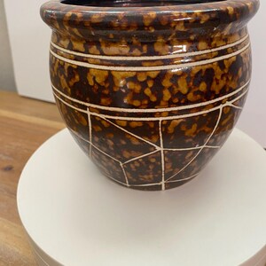May include: A rounded ceramic planter featuring a glossy brown and tan tortoiseshell-like mottled glaze. The exterior is decorated with thin, white incised horizontal lines near the rim and a geometric pattern of intersecting white lines below.