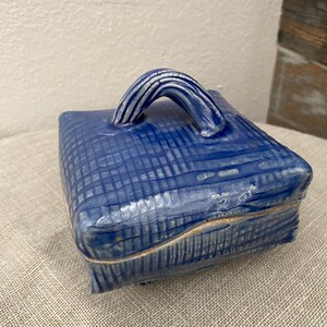 May include: A square, cobalt blue ceramic container with a lid and a curved handle. The container has a textured, crosshatched pattern. The handle is a lighter shade of blue with white accents. The container is likely handmade.