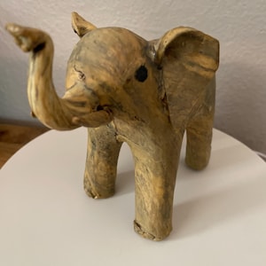 May include: A small, handcrafted elephant figurine with a raised trunk. The elephant is made of a light brown material with darker brown marbling. It has small black eyes and large ears. The figurine is standing on a white surface.