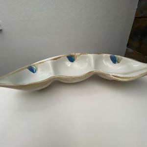 May include: A white ceramic serving dish with three connected compartments. The dish has a wavy, organic shape with a light brown rim and small blue accents. The dish is designed for serving snacks or appetizers.