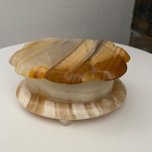May include: A decorative onyx trinket box with a scalloped lid. The box is a mix of beige, brown, and cream colors. The base is round with small feet. The lid has a unique, organic shape. The onyx has a polished, glossy finish.