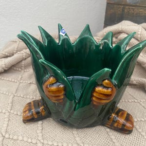 May include: A green ceramic planter shaped like a stylized flower. The planter has a glossy finish and features brown and tan accents on the base and sides. The planter is designed to hold plants or flowers.