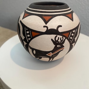 May include: A round ceramic pot featuring a matte white base with hand-painted black and terracotta geometric patterns. The design includes a central black deer figure with a white eye and a terracotta arrow shape on its body, set against a black rim.