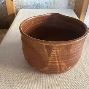 May include: A handmade, brown ceramic bowl with a pouring spout. The bowl has a rounded shape and a textured surface. The interior is a darker brown. The bowl is likely used for cooking or serving.