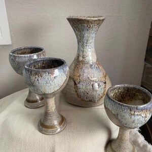May include: A ceramic set consisting of one tall, wide-based pitcher and three stemmed goblets. Each piece features a speckled, mottled glaze in shades of cream, light blue, and earthy brown, with a smooth, glossy finish on the interior and exterior.