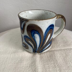 Puede incluir: Una taza de cerámica con una base de color blanco roto moteado que presenta un patrón pintado a mano de formas de pétalos en espiral de color marrón y azul. La taza tiene un asa resistente y un borde redondeado y suave con acabado brillante.