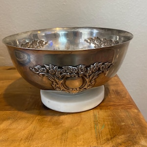 May include: A silver-colored bowl with ornate floral and scrollwork detailing. The bowl sits on a white, circular base. The bowl's rim is slightly flared, and the interior reflects light. The bowl is placed on a wooden surface.