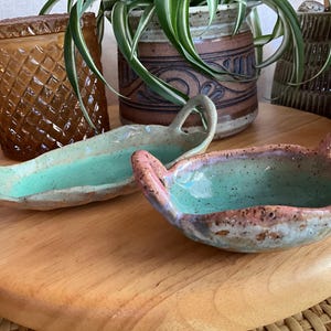 May include: Two handmade ceramic serving dishes with handles. The dishes have a turquoise interior and a speckled, textured exterior with pink and brown accents. They are displayed on a wooden surface, suggesting a rustic or artisanal aesthetic.