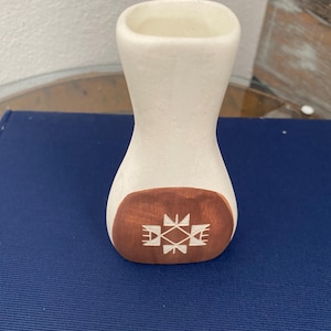 May include: A small, off-white ceramic vase with a flared top and a narrow waist. The base is a dark brown with a white geometric design. The vase is likely handmade and suitable for small flowers or decorative use.
