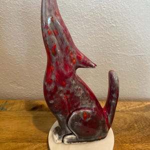 May include: A ceramic wolf figurine in a howling pose. The sculpture has a glossy finish with red, grey, and orange hues. It sits on a round, off-white base, showcasing a detailed and artistic design.