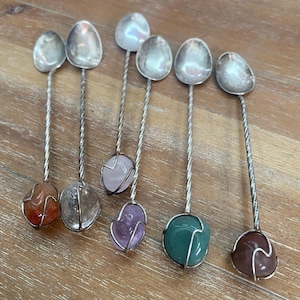 May include: Six silver-toned metal spoons with twisted handles, each featuring a unique polished gemstone at the base. The stones include orange, clear, white, light pink, purple, green, and brown varieties, all secured by thin wire wrapping.