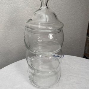 Pode incluir: Um frasco de boticário de vidro transparente com tampa. O frasco é feito de três secções arredondadas empilhadas. A tampa tem uma pega decorativa. O frasco está sobre uma superfície branca.