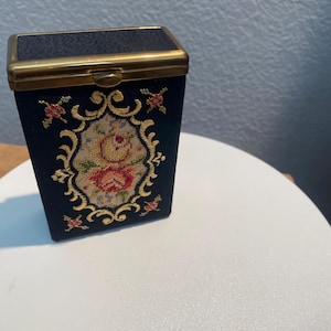May include: A rectangular navy blue fabric case with a gold-toned metal frame and hinged lid. The front features an oval needlepoint embroidery panel depicting a floral arrangement of roses surrounded by a decorative gold-stitched border.
