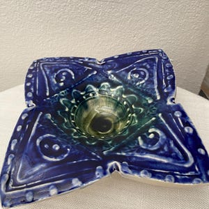 May include: A handcrafted, blue ceramic bowl with a unique, four-lobed design. The bowl features a central well and is adorned with white, raised dot and geometric patterns. The interior has a green and blue glaze, creating a striking contrast.